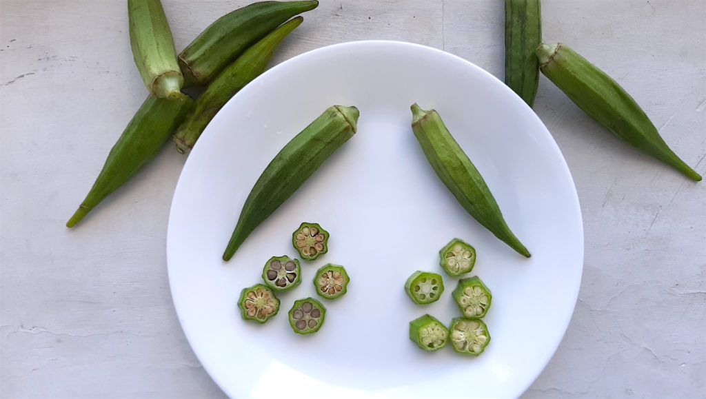 Ripe vs Overripe okra, sliced and whole shown on a white plate with other whole okra in the background. Sugar with Spice Blog