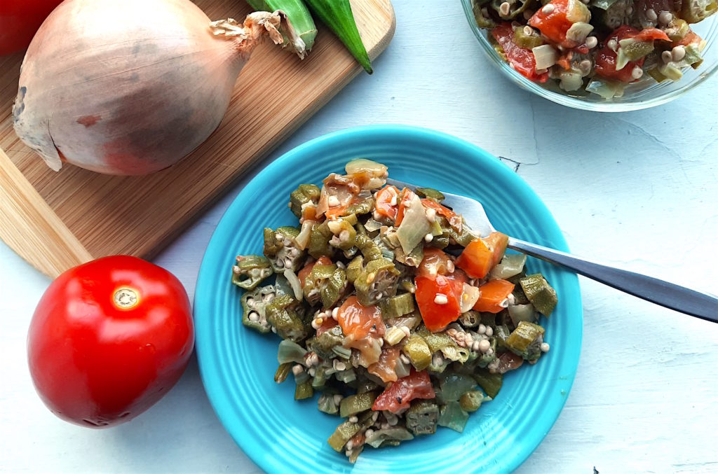 Southern Sautéed okra with onions, garlic, and tomatoes on a blue plate, overhead shot. Glass pyrex bowls with more okra, tomatoes, garlic, and onions in the background. Also a wooden cutting board with a whole onion, whole okra, and tomatoes in the background. Sugar with Spice Blog.