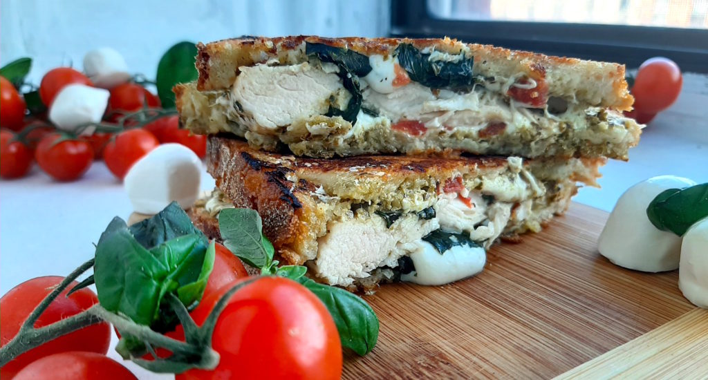 Chicken Caprese Panini with chicken breast, fresh basil, crushed tomatoes, fresh mozzarella on peasant bread. Cut in half and layered on top of each other on a wooden cutting board. Surrounded by cherry tomatoes on the vine, fresh basil and fresh mozzarella balls. On a white window sill. Sugar with Spice Blog.