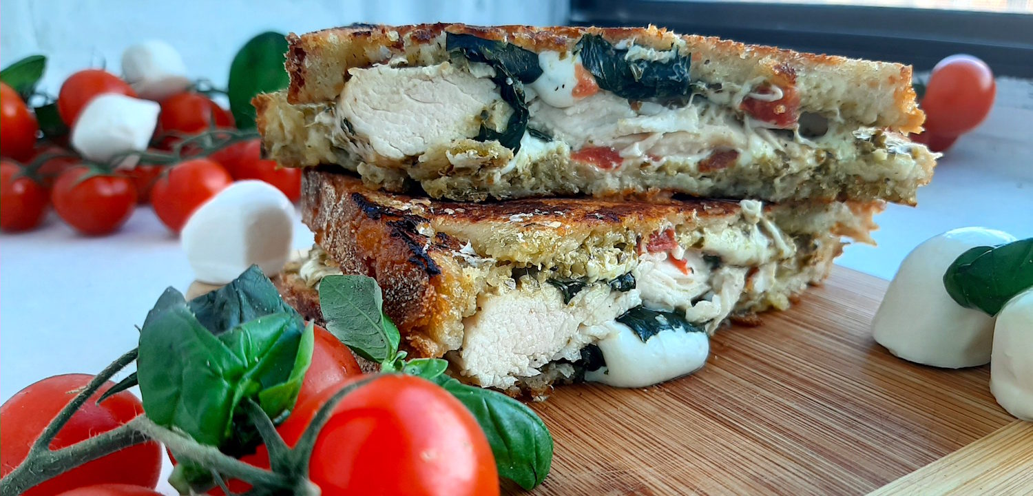 Chicken Caprese Panini with chicken breast, fresh basil, crushed tomatoes, fresh mozzarella on peasant bread. Cut in half and layered on top of each other on a wooden cutting board. Surrounded by cherry tomatoes on the vine, fresh basil and fresh mozzarella balls. On a white window sill. Sugar with Spice Blog.