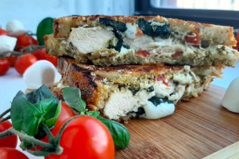 Chicken Caprese Panini with chicken breast, fresh basil, crushed tomatoes, fresh mozzarella on peasant bread. Cut in half and layered on top of each other on a wooden cutting board. Surrounded by cherry tomatoes on the vine, fresh basil and fresh mozzarella balls. On a white window sill. Sugar with Spice Blog.