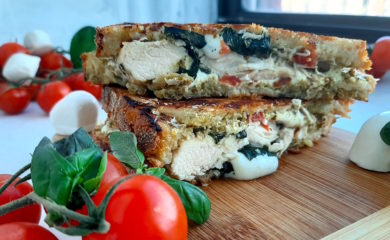 Chicken Caprese Panini with chicken breast, fresh basil, crushed tomatoes, fresh mozzarella on peasant bread. Cut in half and layered on top of each other on a wooden cutting board. Surrounded by cherry tomatoes on the vine, fresh basil and fresh mozzarella balls. On a white window sill. Sugar with Spice Blog.