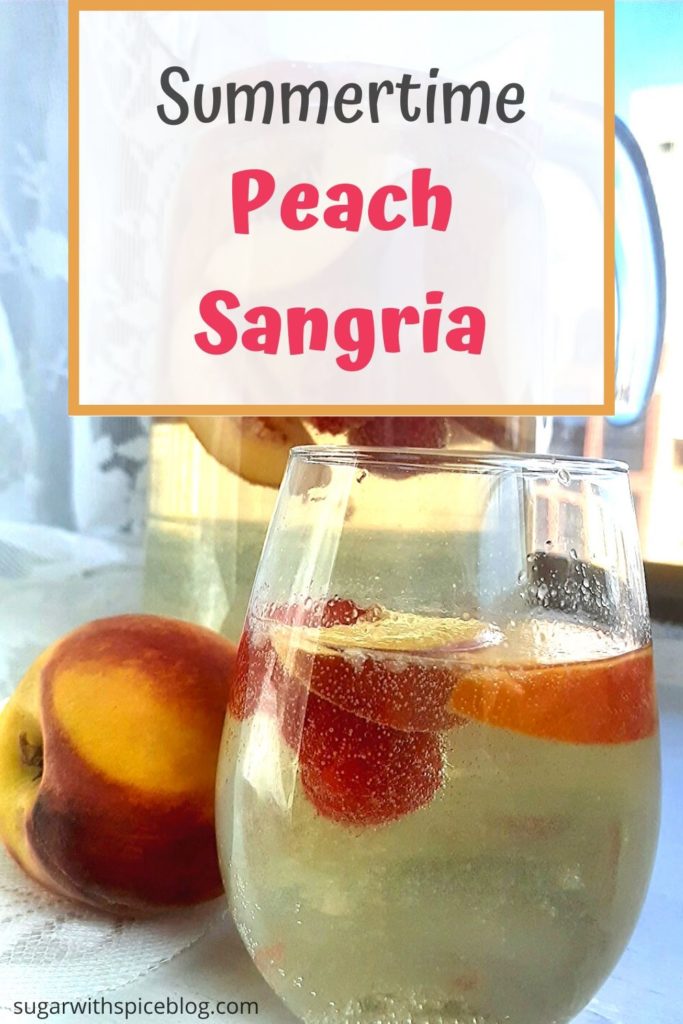 Summertime peach sangria in a pitcher and stemless wine glass on a windowsill with lace curtains, fresh peaches, and fresh raspberries around. Pinterest Image. Sugar with Spice Blog.