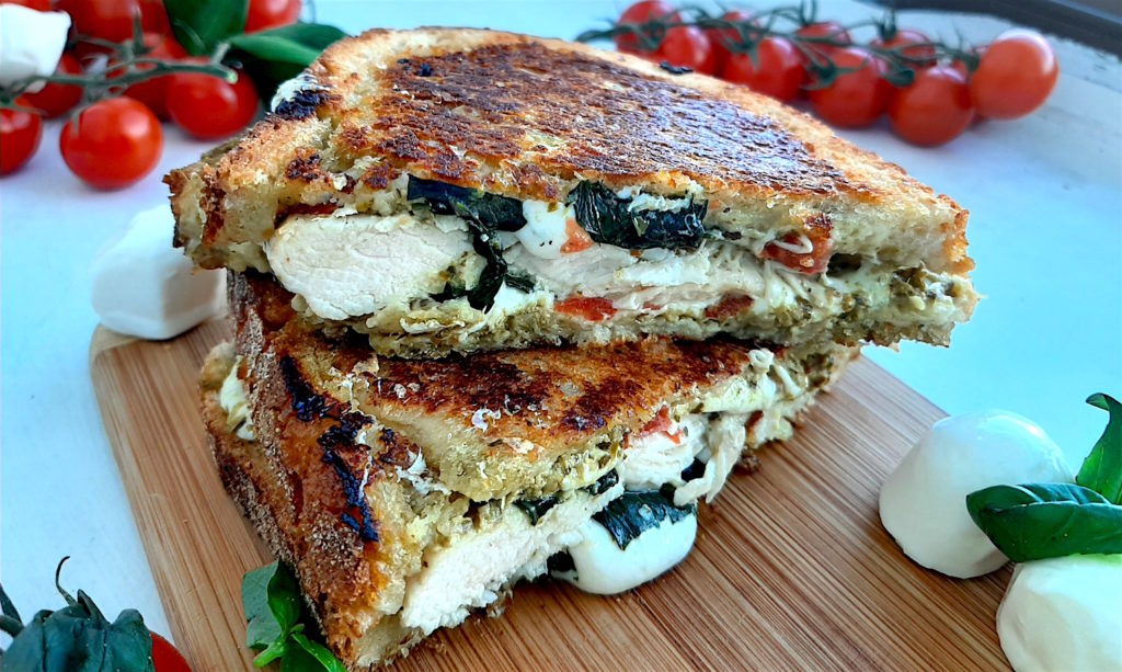 Chicken Caprese Panini with chicken breast, fresh basil, crushed tomatoes, fresh mozzarella on peasant bread. Cut in half and layered on top of each other on a wooden cutting board. Surrounded by cherry tomatoes on the vine, fresh basil and fresh mozzarella balls. On a white window sill. Sugar with Spice Blog.