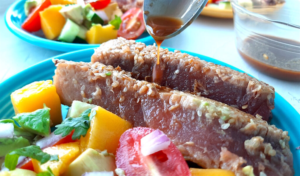 Seared Ahi Tuna slices on a blue plate with avocado, tomato, and mango being drizzled with soy sauce.