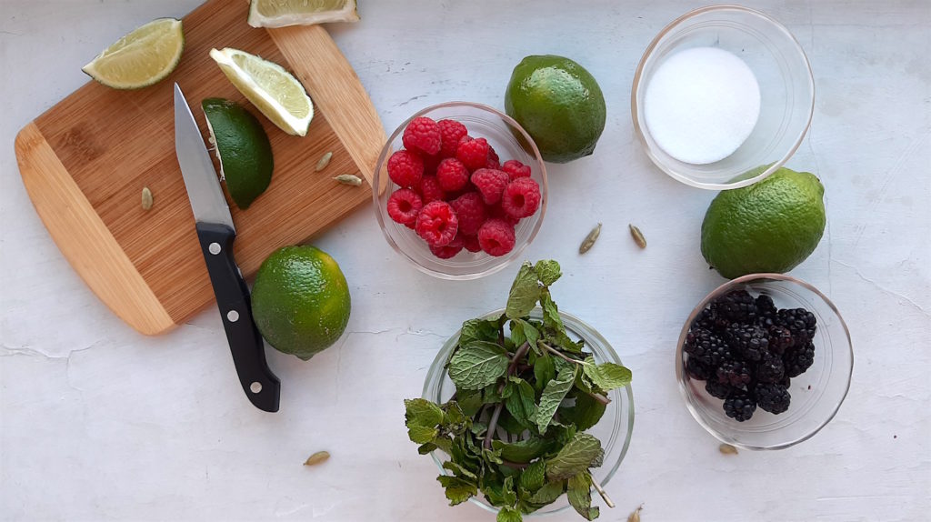 Mixed Berry Mojito Ingredients. Raspberries, Blackberries, Mint Leaves, Sugar in pyrex bowls with a cut lime and knife on a wooden cutting board. Whole limes and cardamom pods scattered around on a white window sill. Overhead shot. Sugar with Spice Blog.