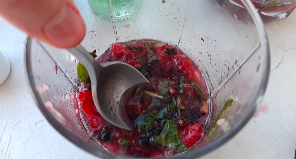 Muddling a Mixed Berry Mojito. Blackberries, raspberries, mint leaves, sugar, ground cardamom, and club soda being muddled with a spoon and a woman's hand.