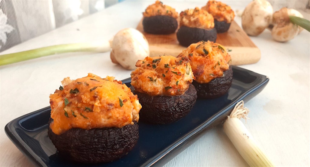 Three Creamy Crab Stuffed Mushrooms on a rectangular blue plate with wooden cutting board and more stuffed mushrooms in the background. Fresh mushrooms and scallions to the side. All on a white window sill. Sugar with Spice Blog.