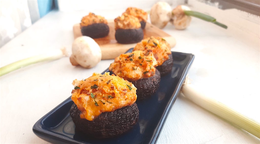 Three Creamy Crab Stuffed Mushrooms on a rectangular blue plate with wooden cutting board and more stuffed mushrooms in the background. Fresh mushrooms and scallions to the side. All on a white window sill. Sugar with Spice Blog.