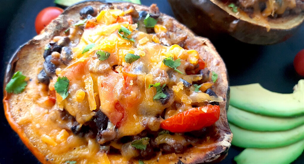 Mexican-style stuffed acorn squash with beans, corn, tomatoes, beef, and cheddar cheese. Two served together in a cast iron skillet with fresh sliced avocado and cherry tomatoes surrounding. Close up shot of one squash. Sugar with Spice Blog.