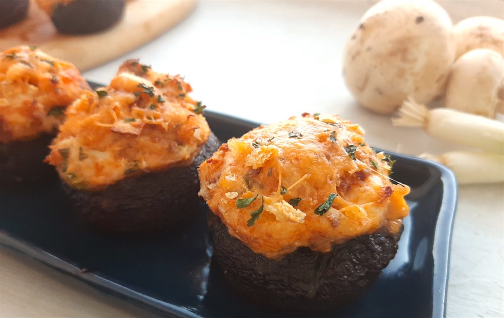 Three Creamy Crab Stuffed Mushrooms on a rectangular blue plate with wooden cutting board and more stuffed mushrooms in the background. Fresh mushrooms and scallions to the side. All on a white window sill. Sugar with Spice Blog.