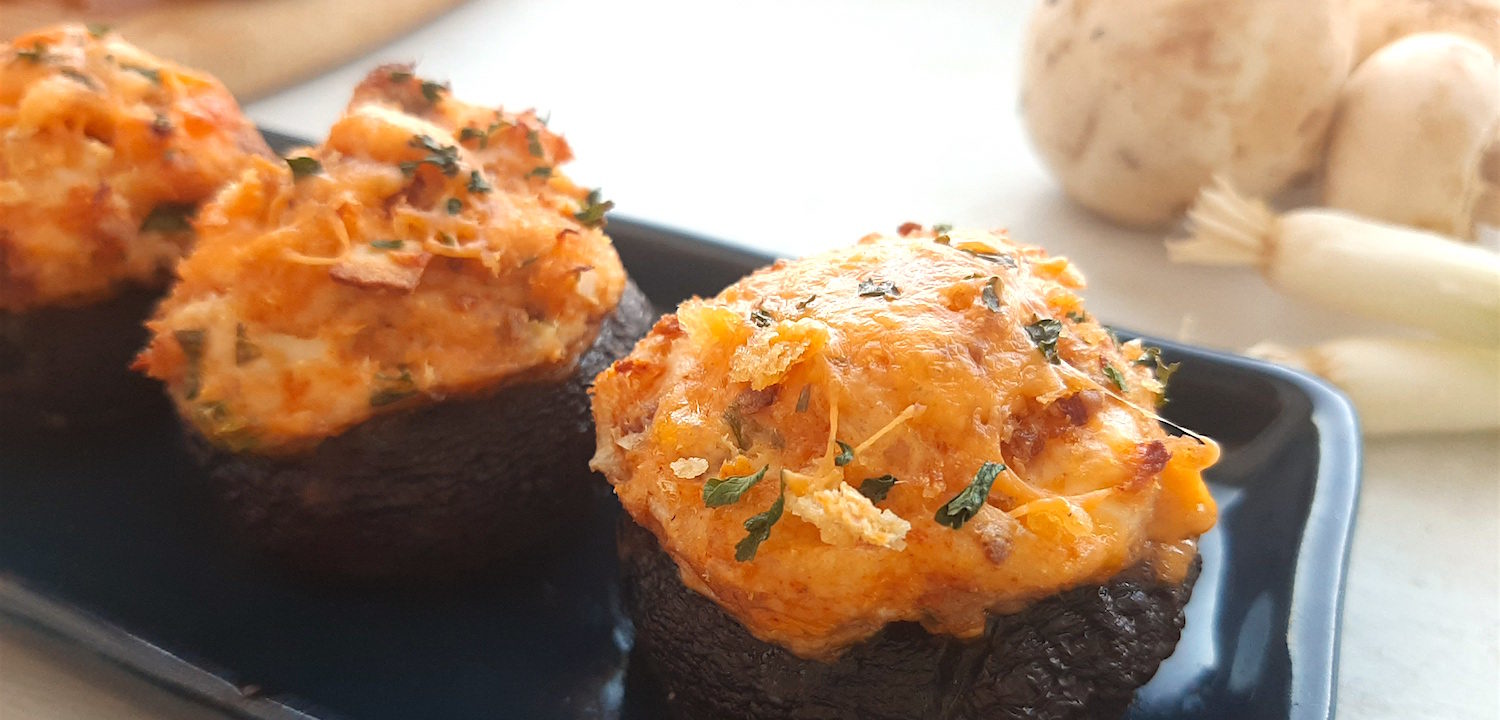 Three Creamy Crab Stuffed Mushrooms on a rectangular blue plate with wooden cutting board and more stuffed mushrooms in the background. Fresh mushrooms and scallions to the side. All on a white window sill. Sugar with Spice Blog.