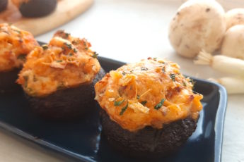 Three Creamy Crab Stuffed Mushrooms on a rectangular blue plate with wooden cutting board and more stuffed mushrooms in the background. Fresh mushrooms and scallions to the side. All on a white window sill. Sugar with Spice Blog.