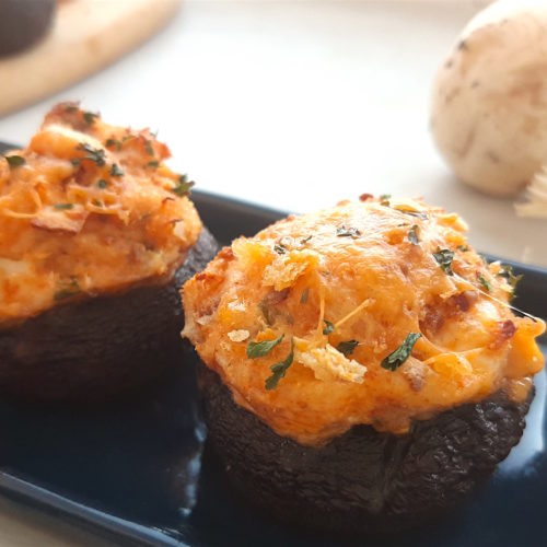 Three Creamy Crab Stuffed Mushrooms on a rectangular blue plate with wooden cutting board and more stuffed mushrooms in the background. Fresh mushrooms and scallions to the side. All on a white window sill. Sugar with Spice Blog.