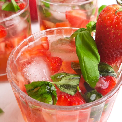 Balsamic Strawberry Basil cocktail, three strawberry basil cocktails in tumblers on a white background. Garnished with fresh basil and strawberries. Surrounded by limes and basil. Sugar with Spice Blog.
