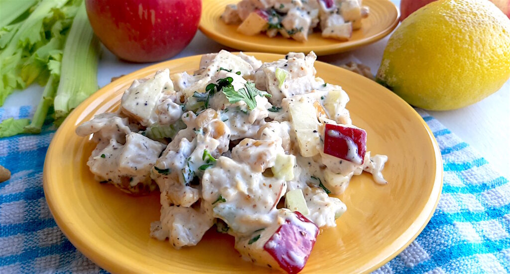 Chicken Salad with Apples and Walnuts served on yellow plates on a blue checked cloth with apples, celery and lemons in the background. Close Up Shot. Sugar with Spice Blog.