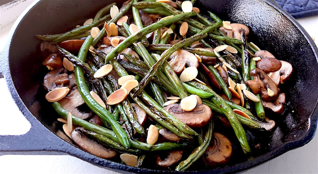 Almond and Mushroom Green Beans in a cast iron skillet surrounded by a blue oven mitt, fresh mushrooms, and sliced almonds. Wide close up shot. Sugar with Spice Blog.