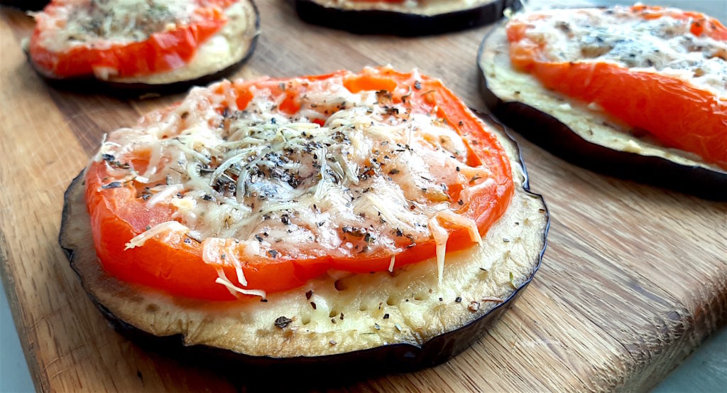 Eggplant pizzas with sliced eggplant layered with sliced tomato, grated parmesan, fresh garlic, and dried parsley. On a wooden cutting board. Sugar with Spice Blog.