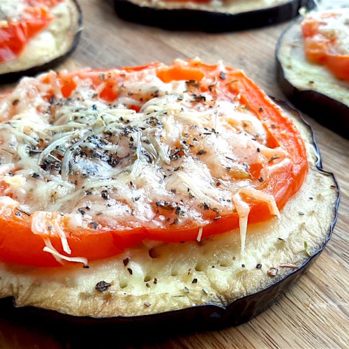 Eggplant pizzas with sliced eggplant layered with sliced tomato, grated parmesan, fresh garlic, and dried parsley. On a wooden cutting board. Sugar with Spice Blog.