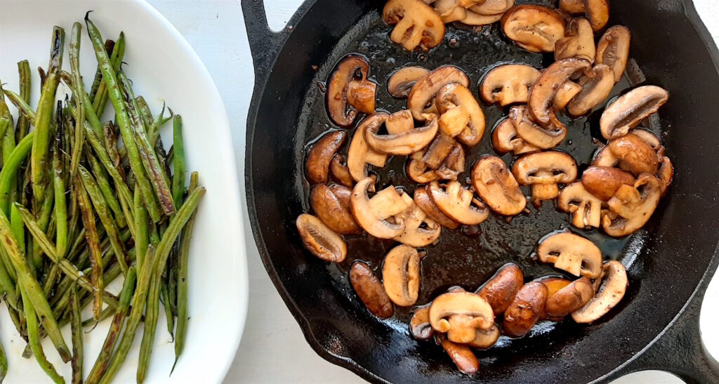Sautéed garlicy mushrooms frying in a cast iron skillet next to fresh green beans ready to combine.