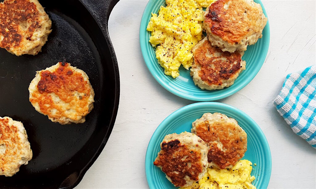 Homemade turkey sausages on a blue plate with creamy scrambled eggs on a white window sill. Next to cast iron skillet with more sausages. Blue checked cloth in the background. Wide shot. Sugar with Spice Blog.