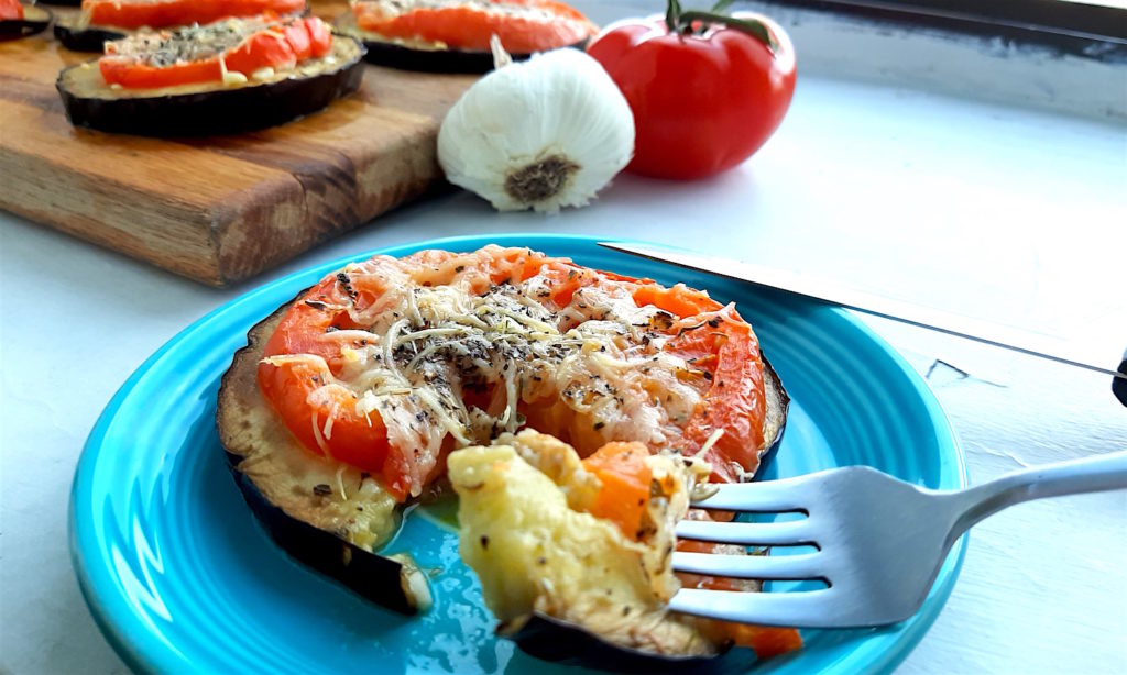 Low-carb eggplant and tomato pizza on a blue plate with one slice cut out on a fork. More eggplant and tomato pizzas in the background on a wooden cutting board surrounded by tomatoes and garlic. Sugar with Spice Blog