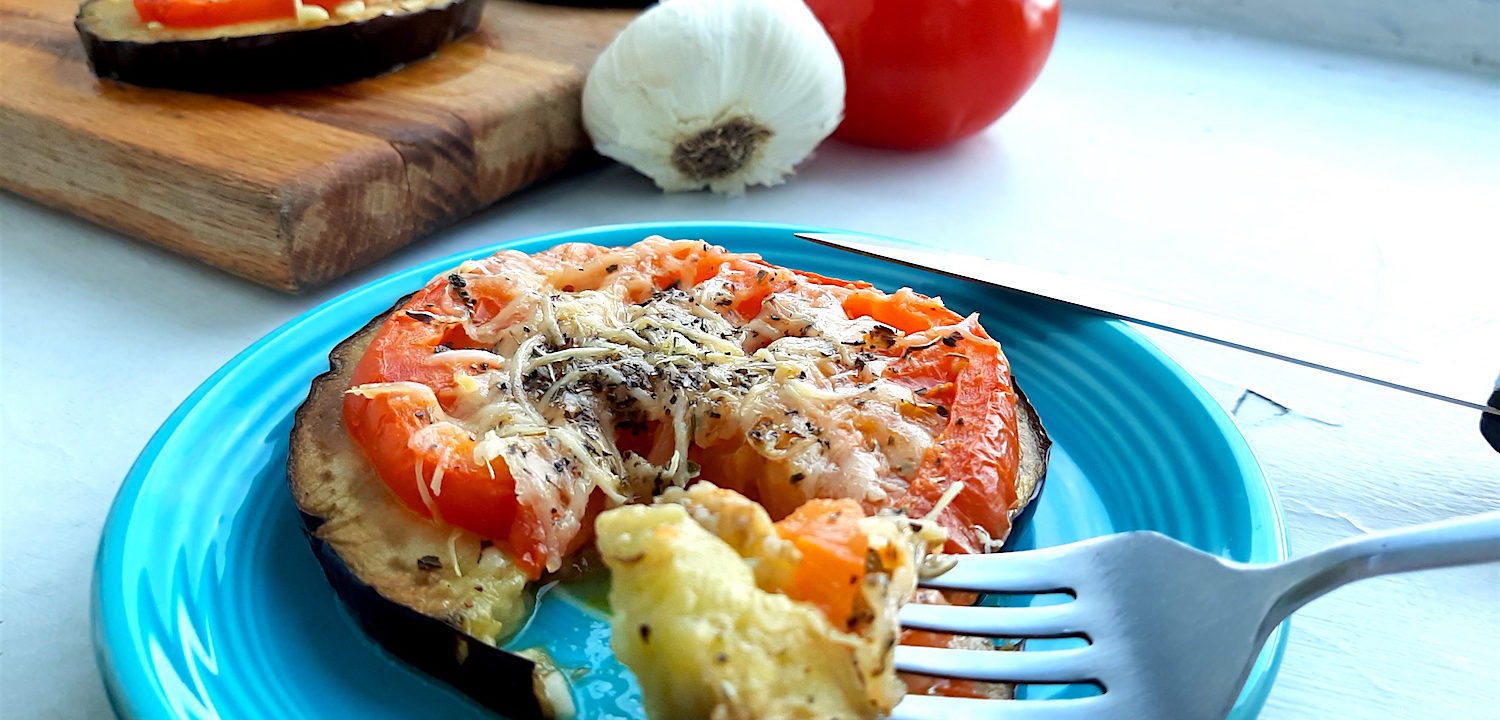 Low-carb eggplant and tomato pizza on a blue plate with one slice cut out on a fork. More eggplant and tomato pizzas in the background on a wooden cutting board surrounded by tomatoes and garlic.