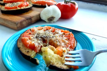 Low-carb eggplant and tomato pizza on a blue plate with one slice cut out on a fork. More eggplant and tomato pizzas in the background on a wooden cutting board surrounded by tomatoes and garlic.
