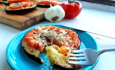 Low-carb eggplant and tomato pizza on a blue plate with one slice cut out on a fork. More eggplant and tomato pizzas in the background on a wooden cutting board surrounded by tomatoes and garlic.
