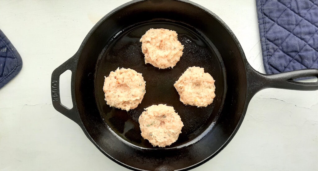 Frying the homemade turkey sausages. Shaped Sausages in a cast iron skillet in oil on a white background with blue oven mitts in the background. Sugar with Spice Blog.