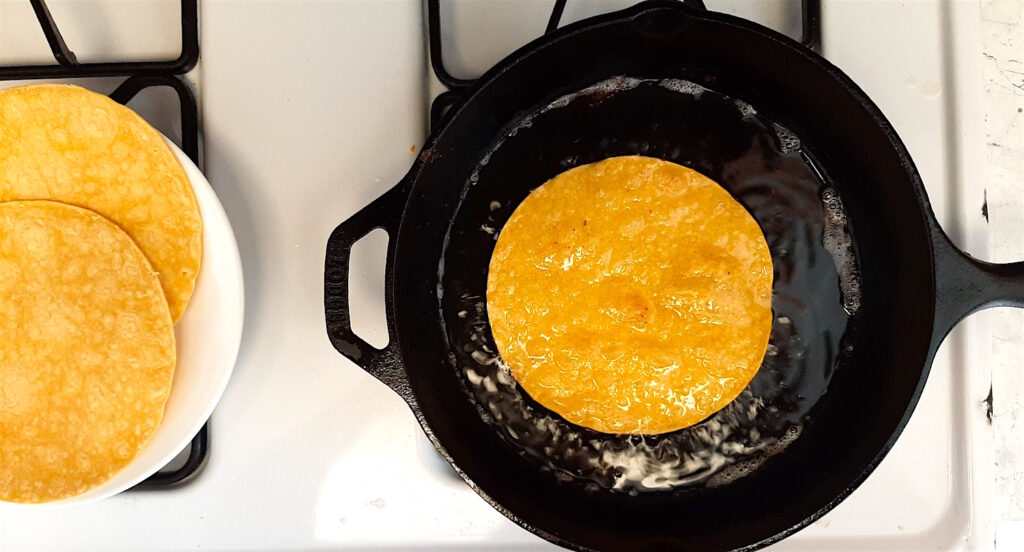 Classic Tex-Mex Migas frying tortillas in bacon lard in black cast iron skillet with more corn tortillas to the side. Sugar with Spice Blog.