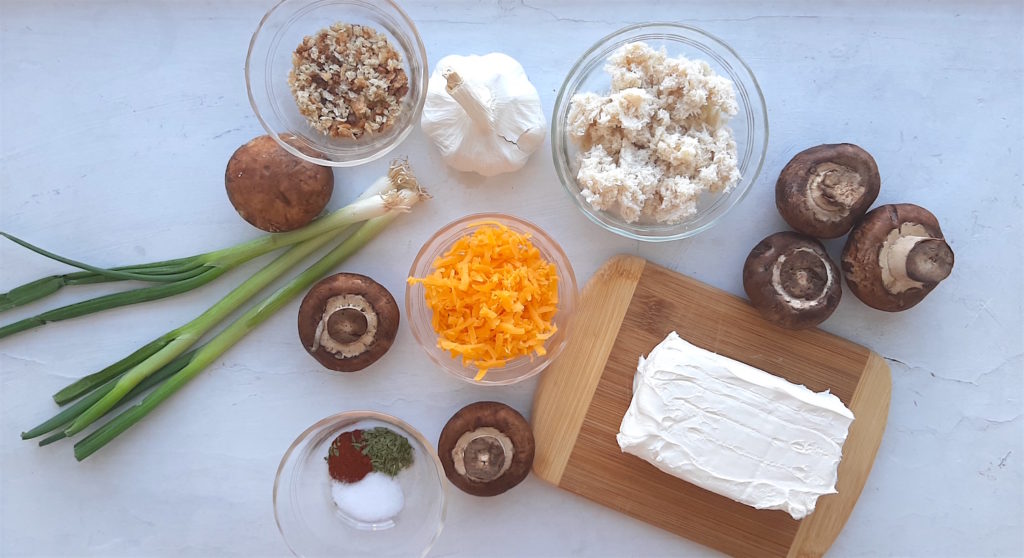 Crab stuffed mushroom ingredients. Grated cheddar, canned crab meat, bread crumbs, paprika, tarragon, and salt in pyrex dishes with scallions, garlic, mushrooms, and cream cheese on a wooden cutting board all on a white window sill, overhead shot. Sugar with Spice Blog.