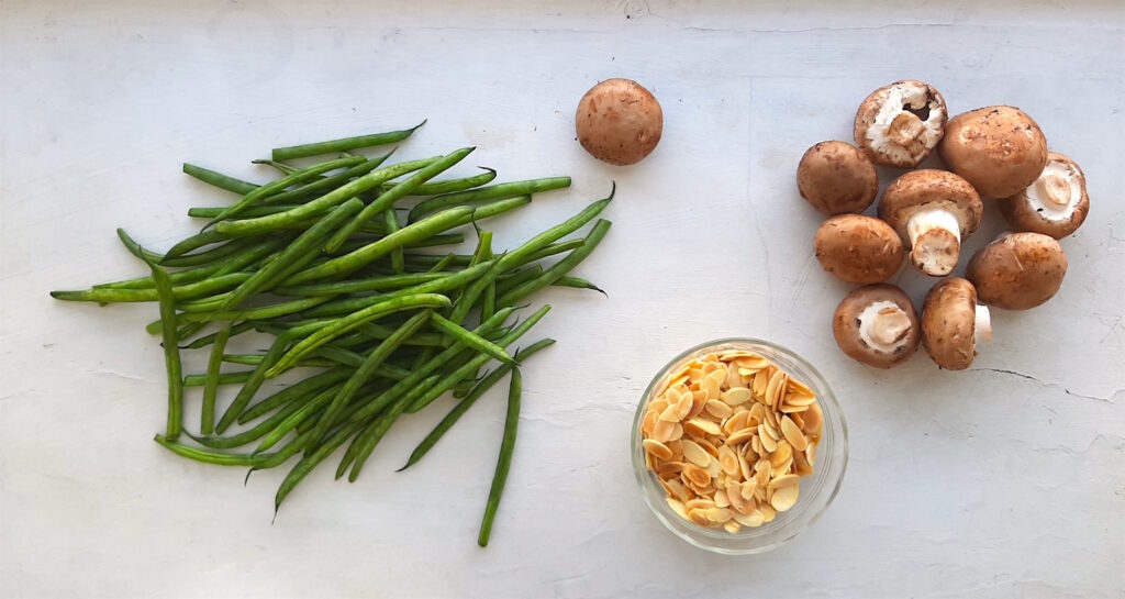 Almond and Mushroom Green Beans ingredients on a white background: fresh green beans, mushrooms, and toasted sliced almonds. Sugar with Spice Blog.