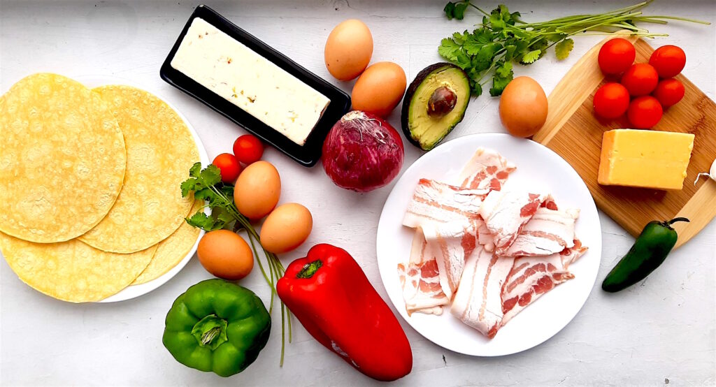 Classic Tex-Mex Migas ingredients. Corn tortillas, red and green bell peppers, eggs, cherry tomatoes, red onion, avocado, fresh cilantro, fresh jalapeño, bacon, cheddar cheese, and pepper jack cheese on a white background. Sugar with Spice blog
