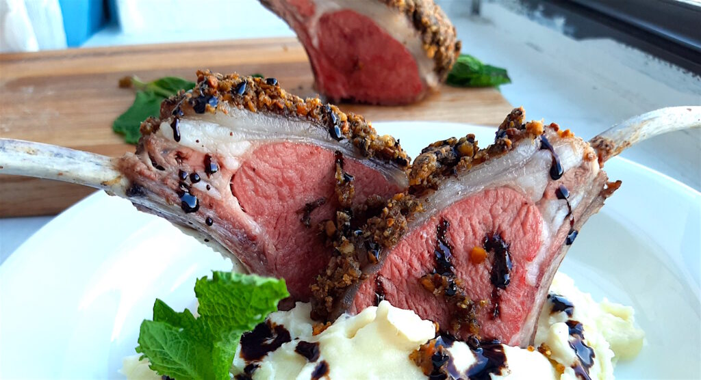 Springtime wine pairing, herb-crusted lamb rack with chianti. Herb crusted lamb rack on a wooden cutting board in the background with two lamb chops in a plate of mashed potatoes with mint leaves and currant glaze in the foreground. Sugar with Spice Blog.
