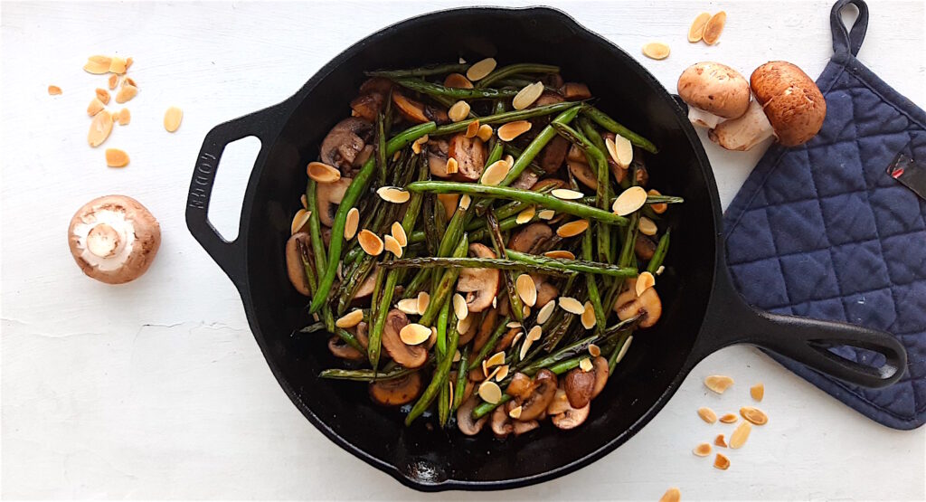 Almond and Mushroom Green Beans in a cast iron skillet surrounded by a blue oven mitt, fresh mushrooms, and sliced almonds. Wide overhead shot. Sugar with Spice Blog.