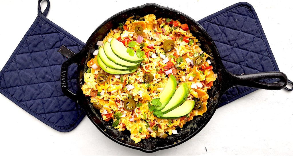 Classic Tex-Mex Migas topped with avocado, pickled jalapeños, and sliced avocado in a cast iron skillet on a white background with blue oven mitts.. Overhead shot. Sugar with Spice Blog.