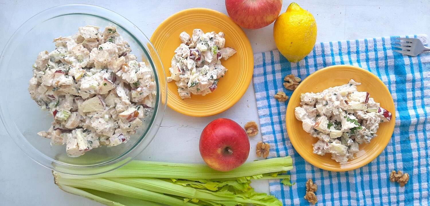 Chicken Salad with Apples and Walnuts served on yellow plates on a blue checked cloth with more salad in a clear pyrex dish nearby, on a white background. Apples, celery, walnuts and lemons around. Wide overhead Shot. Sugar with Spice Blog.