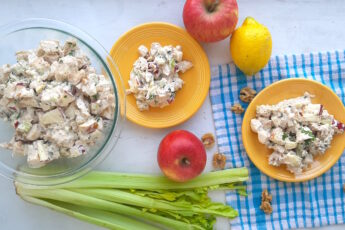 Chicken Salad with Apples and Walnuts served on yellow plates on a blue checked cloth with more salad in a clear pyrex dish nearby, on a white background. Apples, celery, walnuts and lemons around. Wide overhead Shot. Sugar with Spice Blog.