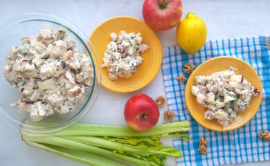Chicken Salad with Apples and Walnuts served on yellow plates on a blue checked cloth with more salad in a clear pyrex dish nearby, on a white background. Apples, celery, walnuts and lemons around. Wide overhead Shot. Sugar with Spice Blog.