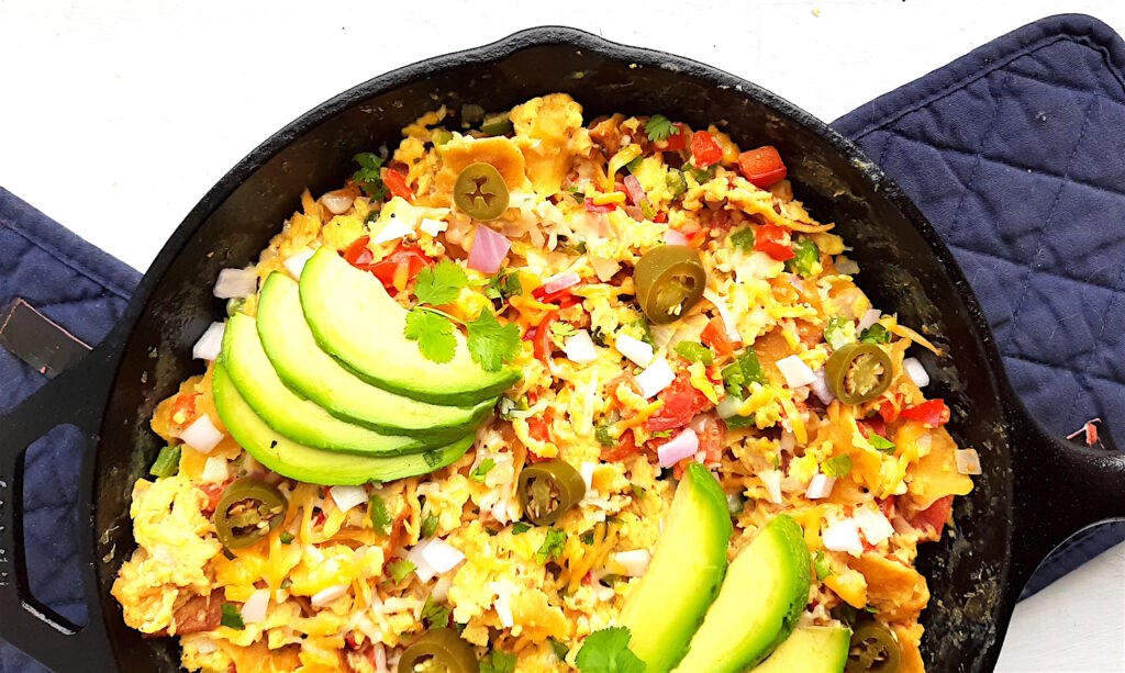 Classic Tex-Mex Migas topped with avocado, pickled jalapeños, and sliced avocado in a cast iron skillet on a white background with blue oven mitts.. Overhead off kilter shot. Sugar with Spice Blog.