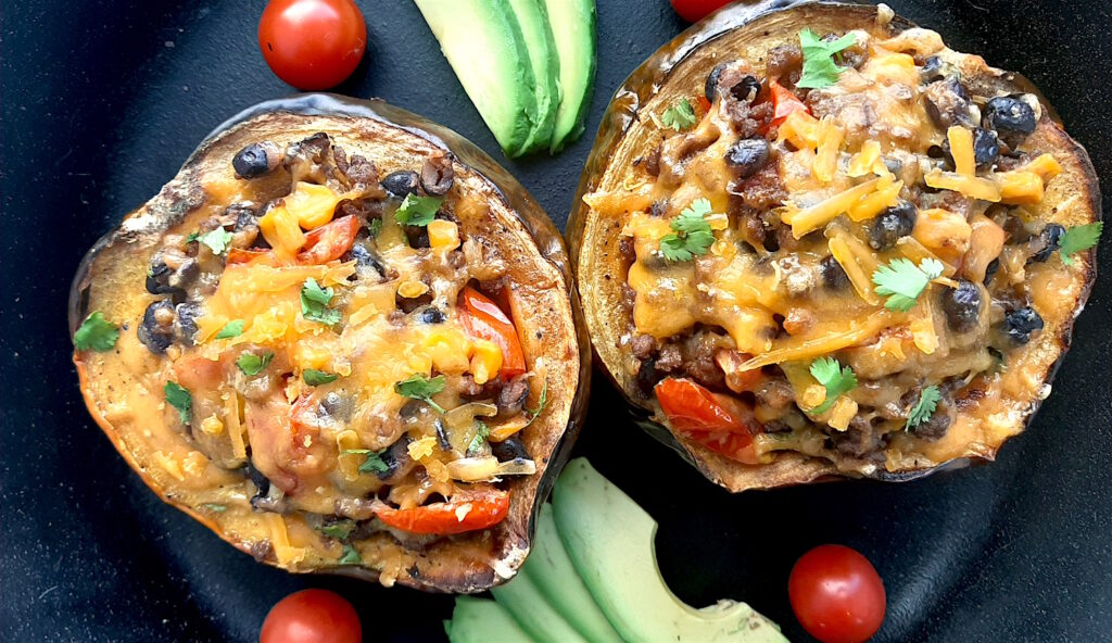 Mexican-style stuffed acorn squash with beans, corn, tomatoes, beef, and cheddar cheese. Two served together in a cast iron skillet with fresh sliced avocado and cherry tomatoes surrounding. Overhead shot. Sugar with Spice Blog.
