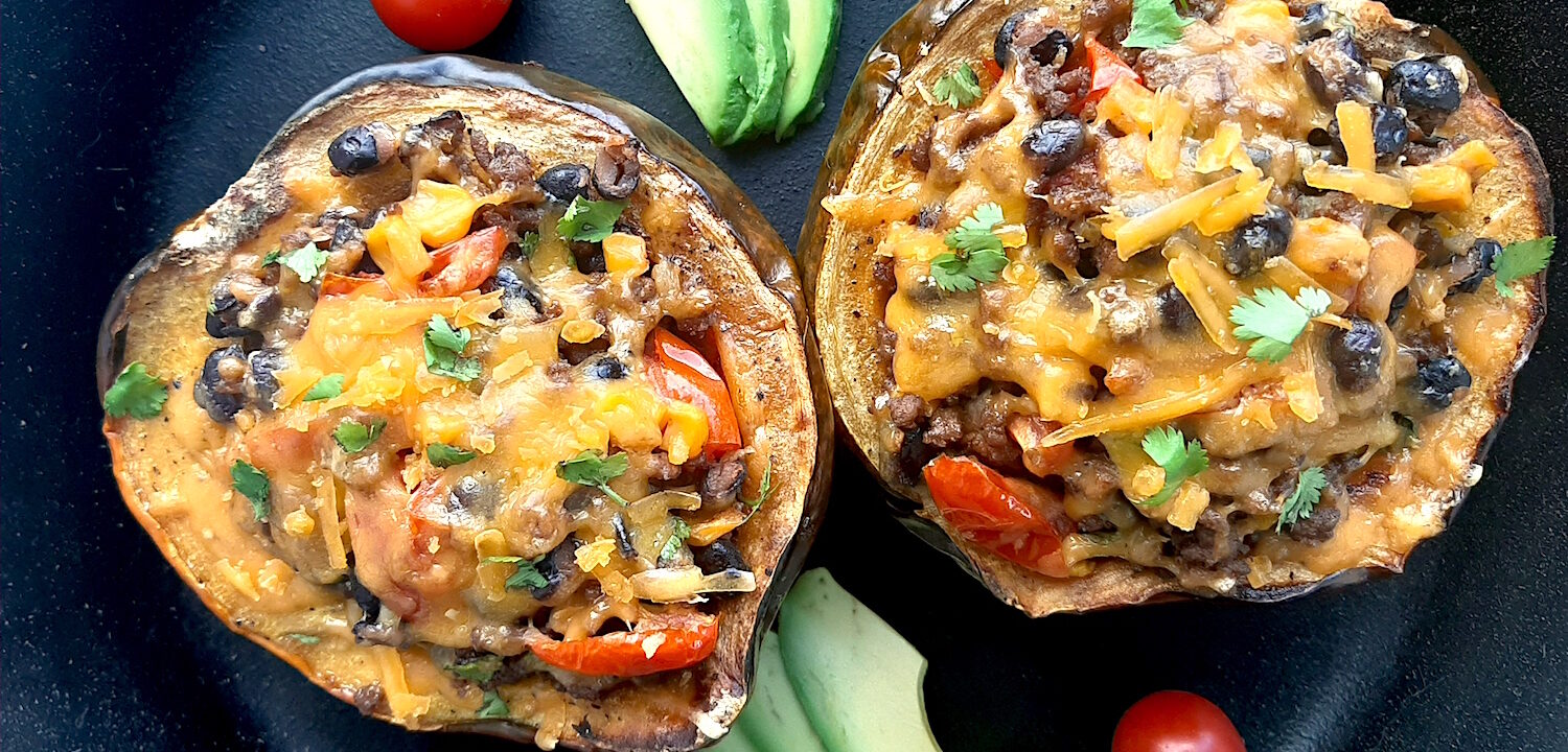 Mexican-style stuffed acorn squash with beans, corn, tomatoes, beef, and cheddar cheese. Two served together in a cast iron skillet with fresh sliced avocado and cherry tomatoes surrounding. Overhead shot. Sugar with Spice Blog.