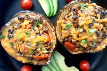 Mexican-style stuffed acorn squash with beans, corn, tomatoes, beef, and cheddar cheese. Two served together in a cast iron skillet with fresh sliced avocado and cherry tomatoes surrounding. Overhead shot. Sugar with Spice Blog.