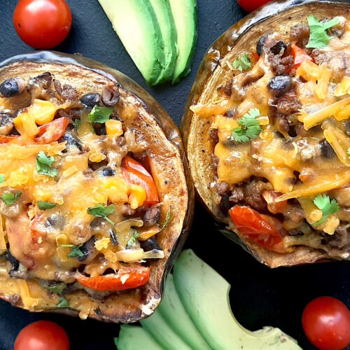 Mexican-style stuffed acorn squash with beans, corn, tomatoes, beef, and cheddar cheese. Two served together in a cast iron skillet with fresh sliced avocado and cherry tomatoes surrounding. Overhead shot. Sugar with Spice Blog.