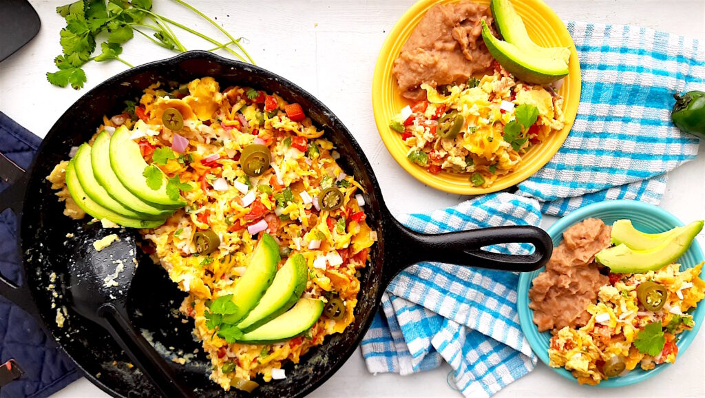 Classic Tex-Mex Migas in a cast iron skillet, a blue plate with refried beans, and a yellow plate with refried beans. All topped with fresh avocado and jalapeños. Blue checked cloth, cilantro, blue oven mitts, and jalapeños surround. Overhead wide shot. Sugar with Spice Blog.