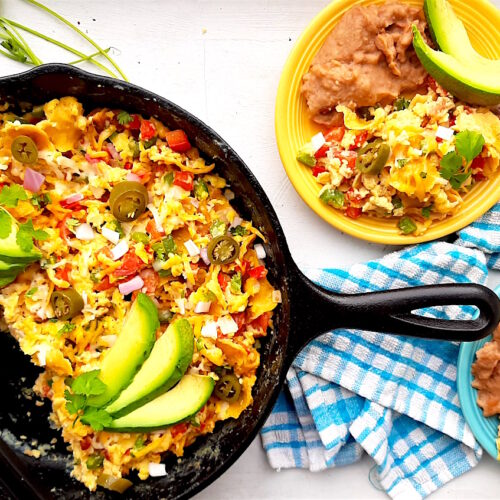 Classic Tex-Mex Migas in a cast iron skillet, a blue plate with refried beans, and a yellow plate with refried beans. All topped with fresh avocado and jalapeños. Blue checked cloth, cilantro, blue oven mitts, and jalapeños surround. Overhead wide shot. Sugar with Spice Blog.