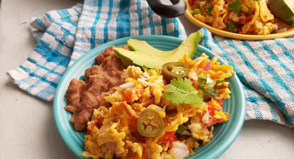 Classic Tex-Mex Migas a blue plate with refried beans, and a yellow plate with refried beans. All topped with fresh avocado and jalapeños. Blue checked cloth, cilantro, and jalapeños surround. Wide close up shot. Sugar with Spice Blog.