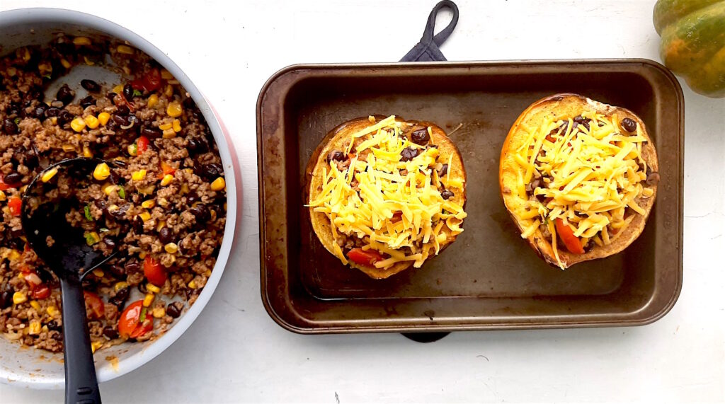 Stuffed and filled Mexican-Style Stuffed Acorn Squash. Two halves of filled squash in a metal baking dish. Pan of filling with black serving spoon near by. Whole acorn squash and blue oven mitts nearby. White background. Sugar with Spice Blog.