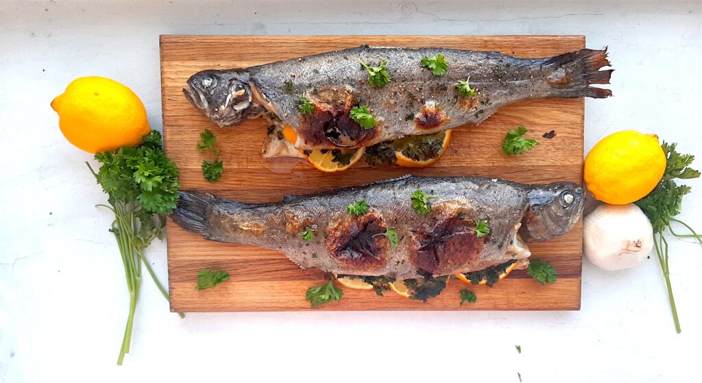 Whole roasted rainbow trout on a wooden cutting board with fresh lemons and parsley surrounding on a white background. Sugar with Spice Blog.